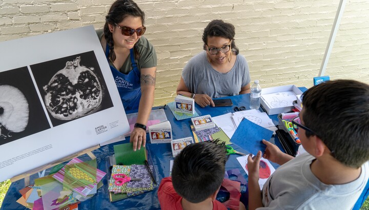 Two adults sit behind a table showing two children art supplies.