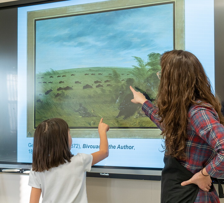 A child stands in front of a screen and points to a detail in an artwork on the screen; an adult standing next to the child points out a different detail