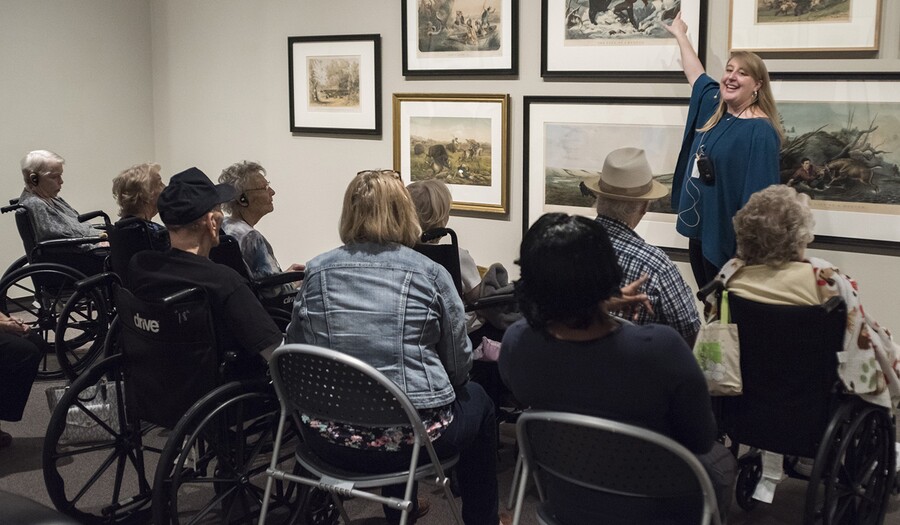 A group of senior citizens sit in chairs and wheelchairs around a woman who is gesturing toward framed artwork on a wall.