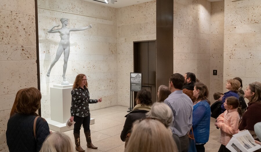 A group of visitors stand around a woman who is talking in front of a large sculpture.