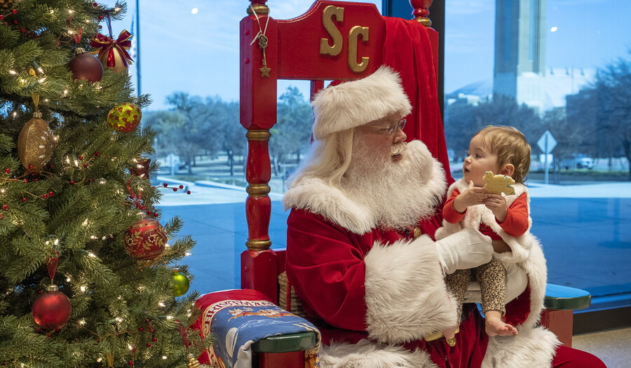 Santa Claus looks at a toddler he holds in his arms.
