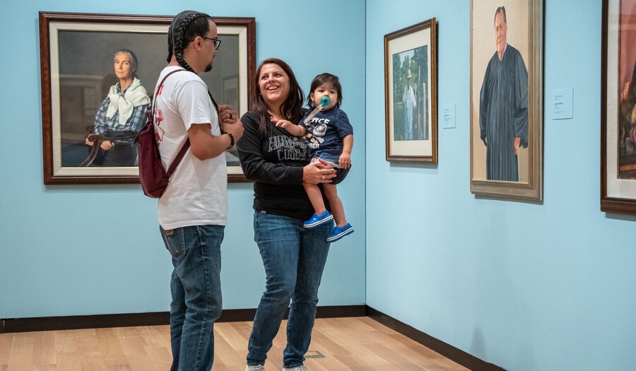 Three people in a gallery; the person on the right holds a child in her arms and looks with a smile at the person on the left